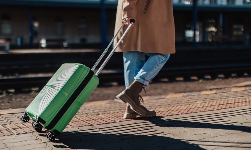 A person in a tan coat and jeans rolls a green suitcase on a train platform