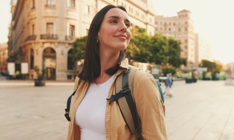 Smiling woman with backpack walks through a sunlit city square, surrounded by historic buildings and greenery